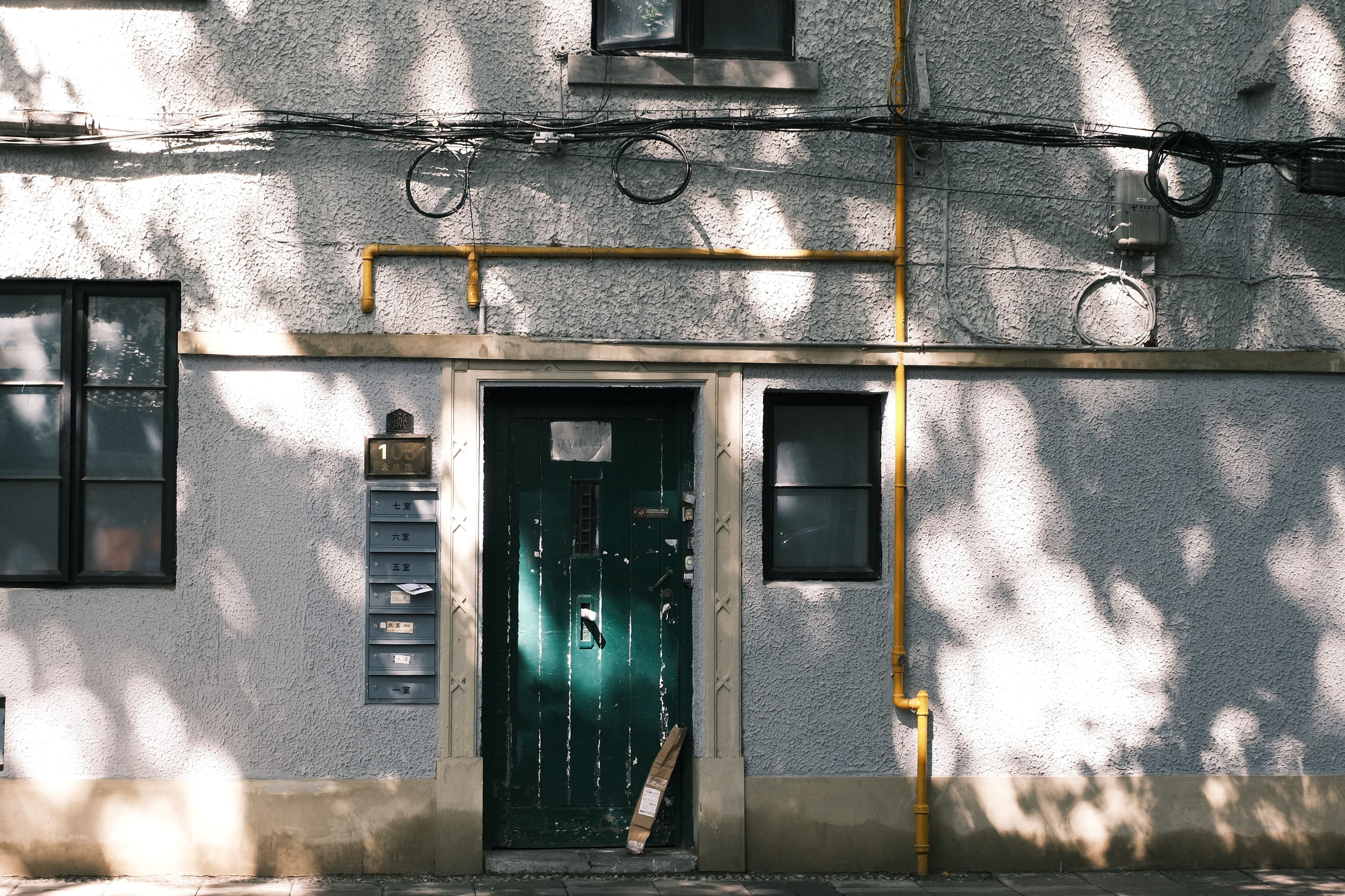 Exterior wall with green door, shadows, and urban elements including pipes and wires.