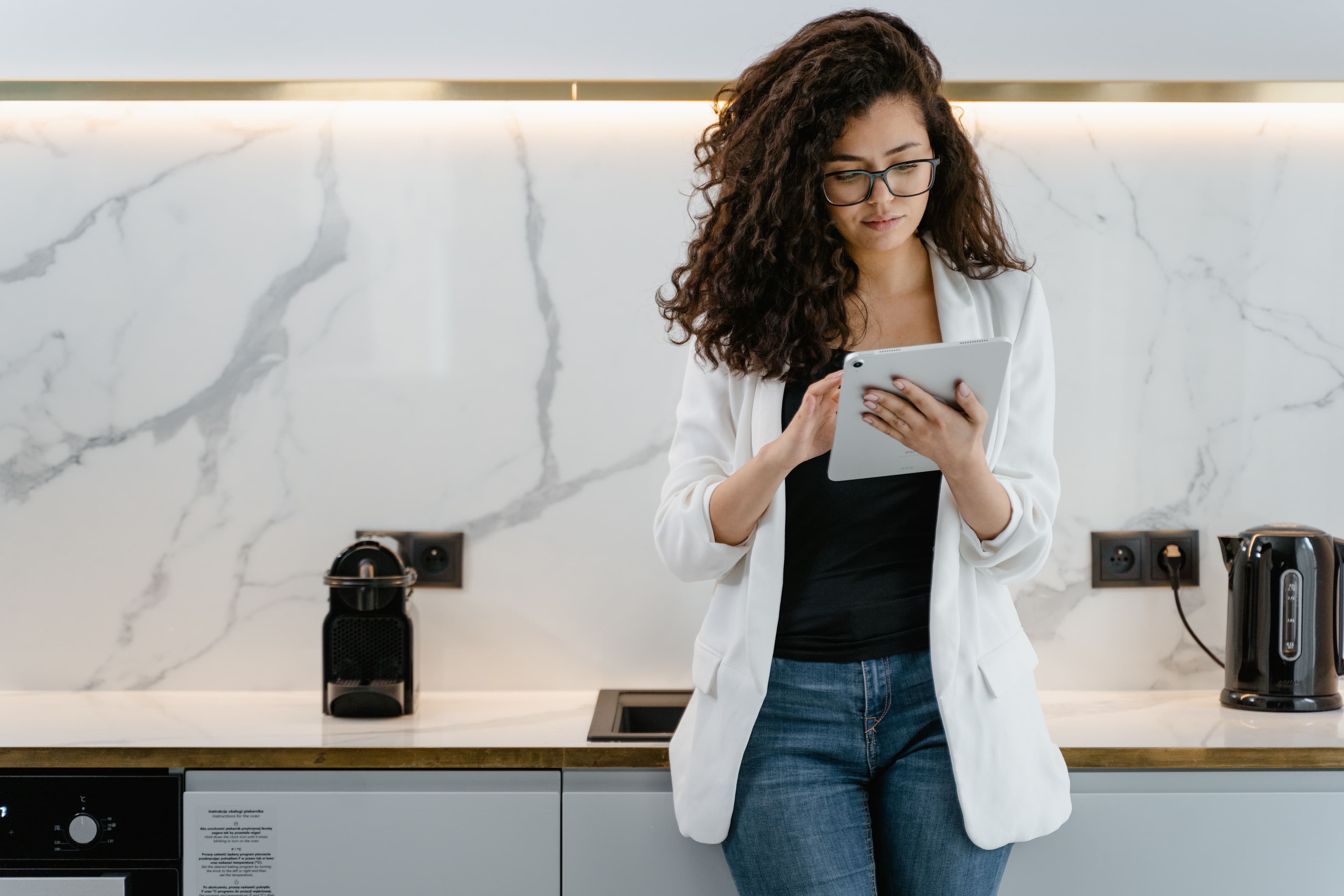 Woman in a white blazer using a tablet in a contemporary kitchen setting.