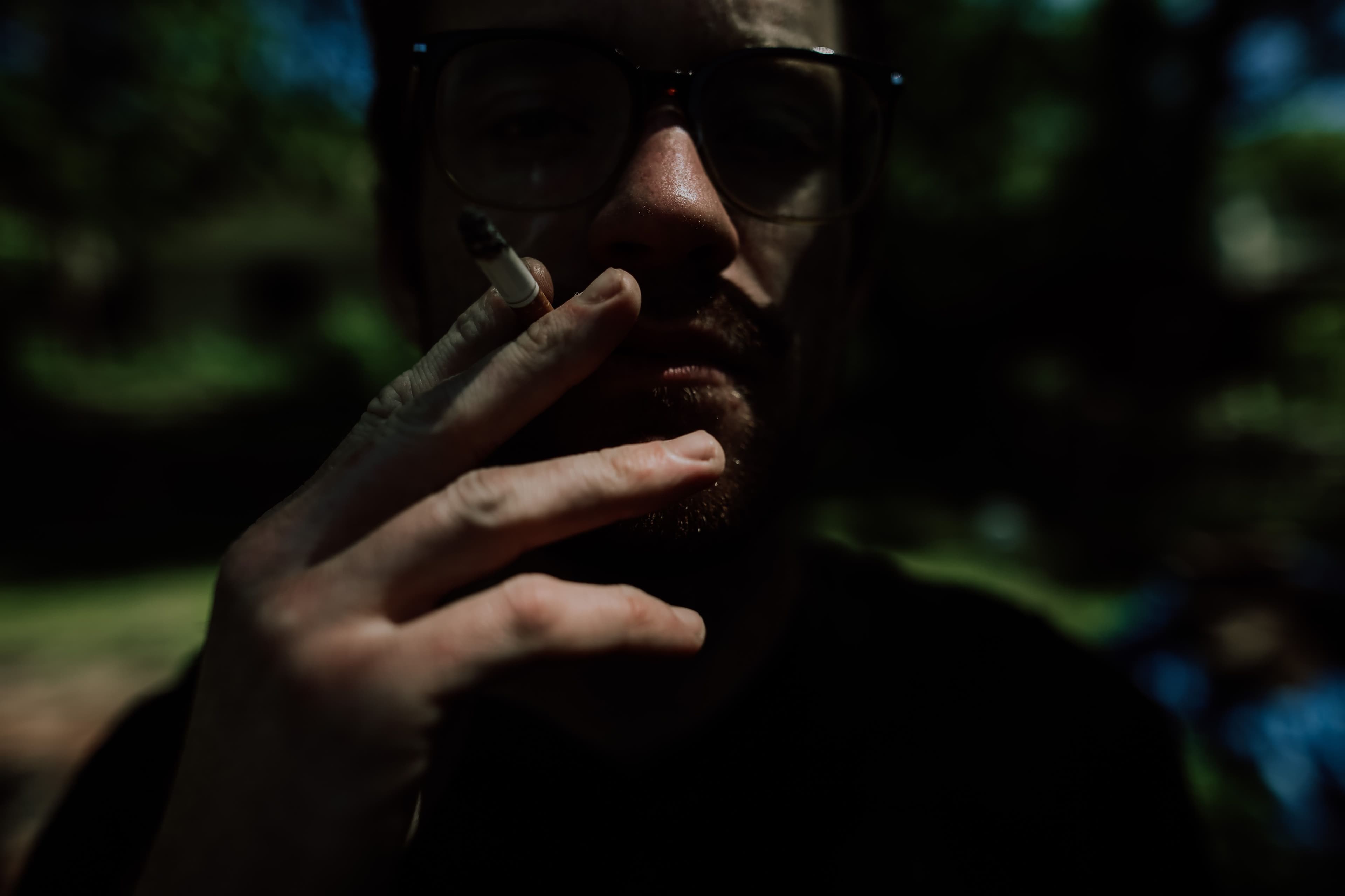 Unrecognizable serious male in eyeglasses standing on street in sunny summer day and smoking in shadow