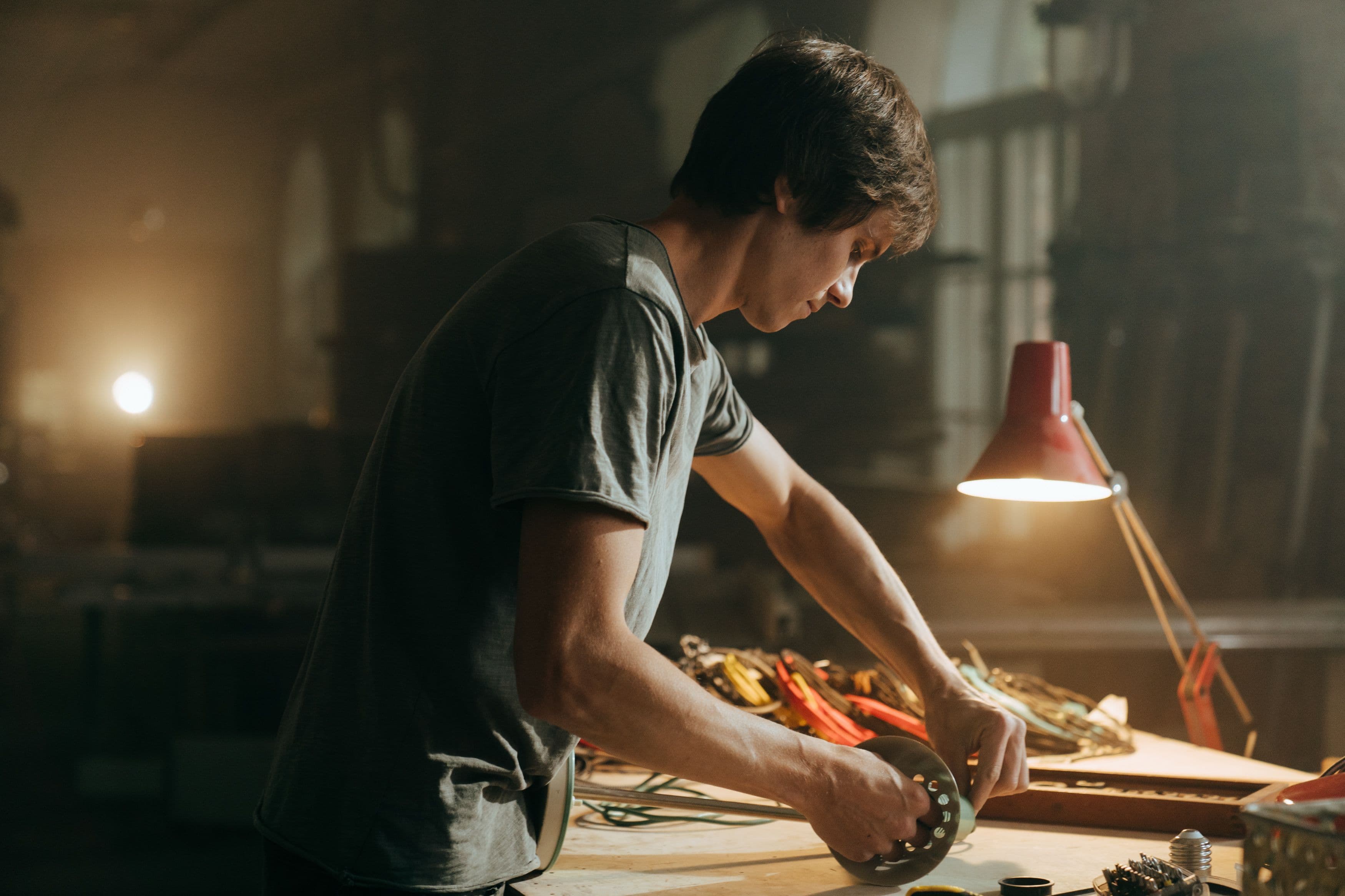 Craftsman focusing on his work at a cluttered bench in a dimly lit workshop, illuminated by a desk lamp.