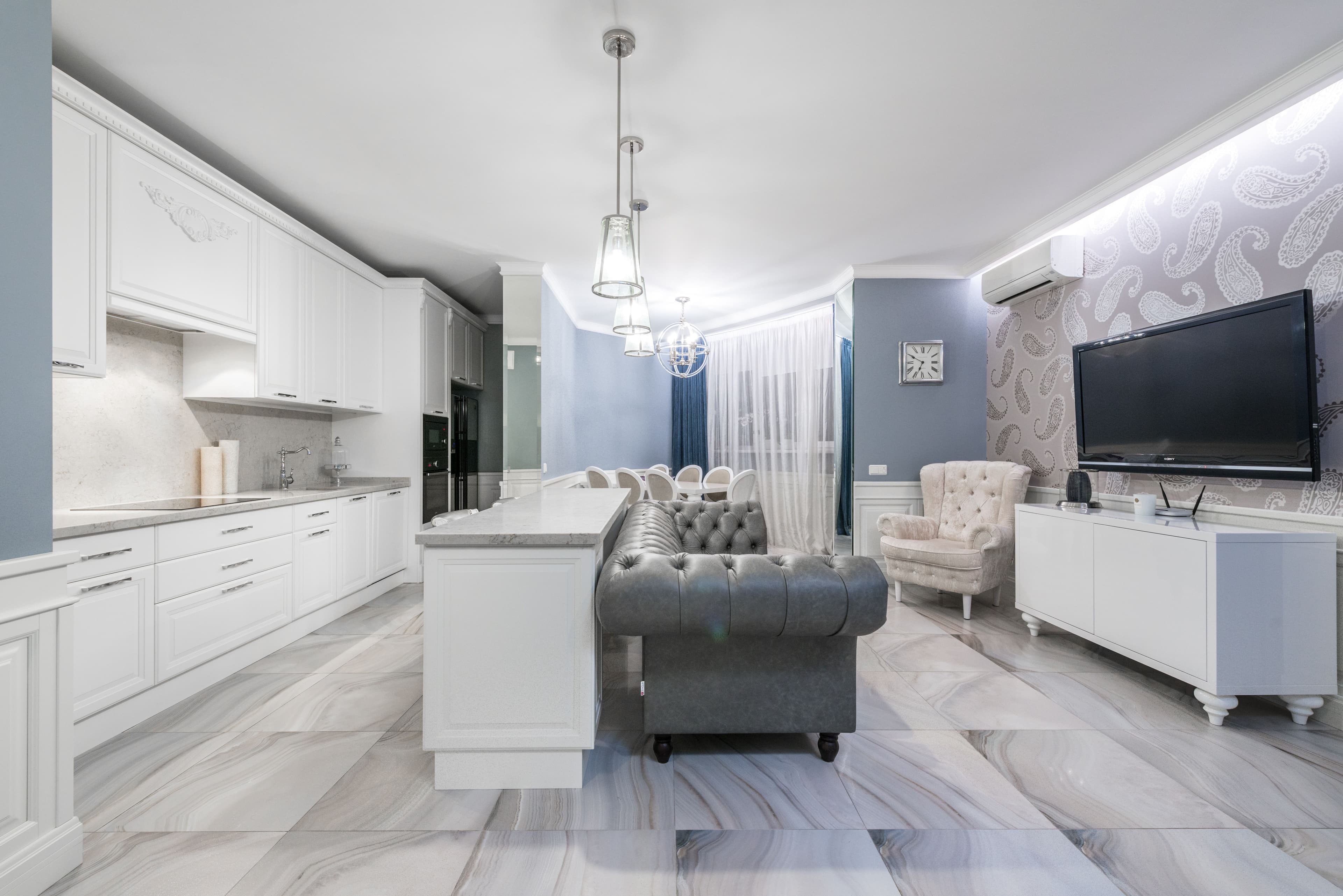 Contemporary interior of dining zone with white cabinets and island counter connected with living room with couch and TV on wall
