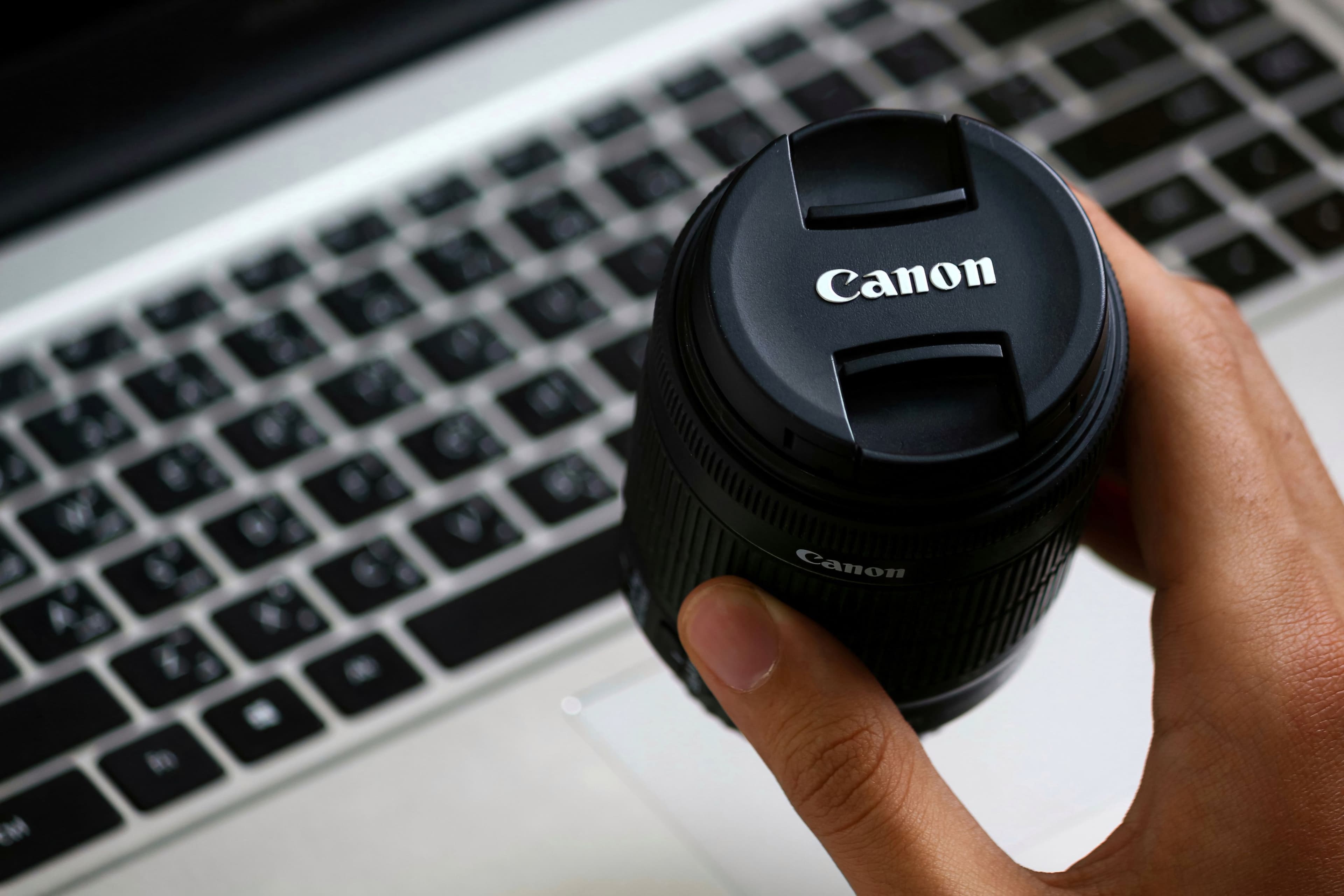 Hand holding Canon lens above a laptop, showcasing modern photography gear.